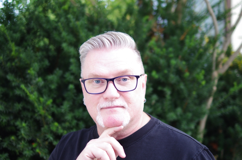 Portrait of David Moulton Counsellor for Toronto wearing blue glasses with a curious expression standing in front of a Japanese yew bush