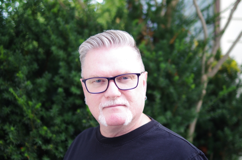 Portrait of David Moulton Counsellor for Toronto wearing blue glasses standing in front of a Japanese Yew bush
